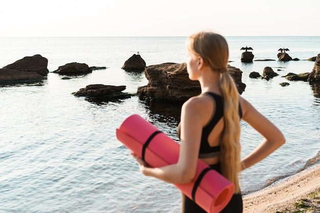 sportliche konzentrierte Frau, die einen Trainingsanzug trägt, der das Meer betrachtet und die Yogamatte hält, die am Morgen im Freien arbeitet