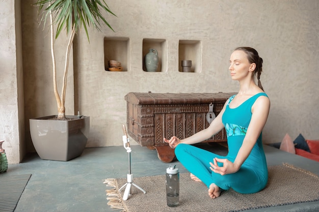 Sportliche schlanke Frau in Sportbekleidung meditieren in Lotussitz mit Telefon und Flasche Wasser
