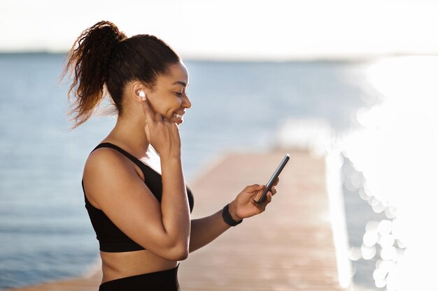 Sportliche schwarze Frau, die auf den Ohrstöpsel drückt und auf den Smartphone-Bildschirm schaut