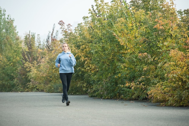 Sportliche und gesunde junge blonde Frau in Activewear, die morgens im Herbst im Park zwischen grünen und gelben Bäumen joggt