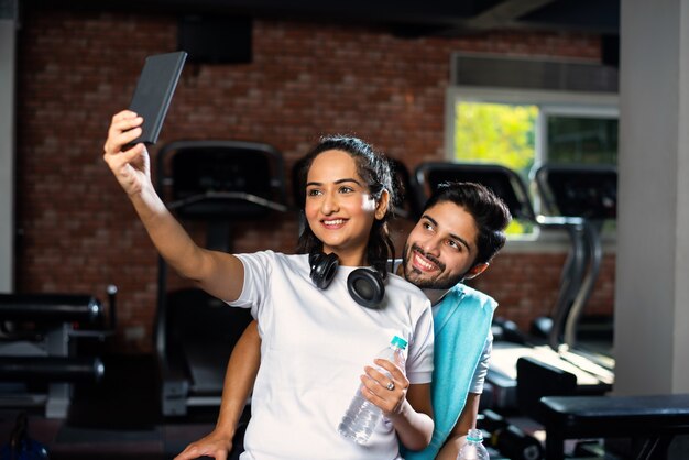 Sportlicher indischer asiatischer Mann und Frau, die sich zwischen den Übungen ausruhen und das Smartphone im Fitnessstudio benutzen, etwas präsentieren oder ein Selfie-Foto machen