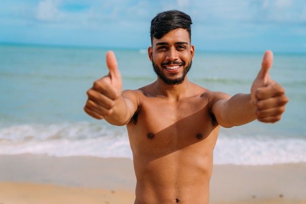 Sportlicher junger lateinamerikanischer Mann am Strand mit Daumen nach oben