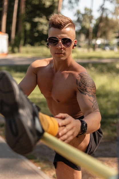 Sportlicher junger Mann mit Frisur, Sonnenbrille und Kopfhörern in schwarzen Turnschuhen und Shorts beim Stretching und Training im Park