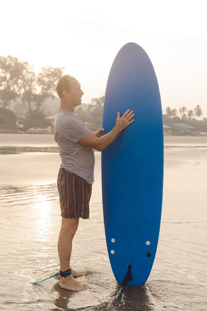 Sportlicher Mann mit am Knöchel befestigter Leine, die Hand auf dem Surfbrett haltend und die Ausrüstung auf dem Sand betrachtend