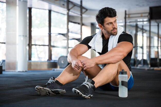 Sportlicher Mann mit blauen Shorts und schwarzem T-Shirt, der im Fitnessstudio auf dem Boden sitzt