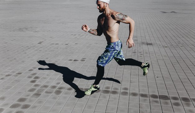 Sportlicher Mann mit nacktem Oberkörper mit Tätowierungen und Stirnband auf dem Kopf in schwarzen Leggings und blauen Shorts läuft an einem warmen sonnigen Tag auf Pflastersteinen.