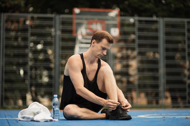 Sportlicher muskulöser Mann, der vor dem morgendlichen Lauf auf dem Basketballplatz im Freien Schnürsenkel an Sportturnschuhen bindet