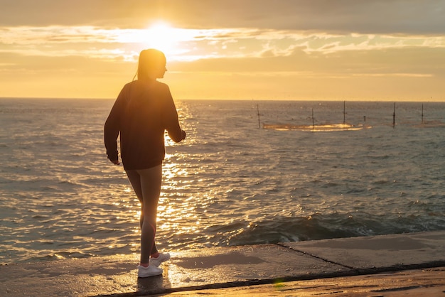 Sportliches athletisches Mädchen ruht sich nach einem Lauf am Meer bei Sonnenuntergang aus, führt einen aktiven Lebensstil