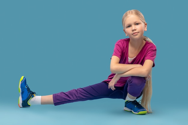 Sportliches Aufwärmen. Selbstbewusstes süßes Mädchen in Sportkleidung und Turnschuhen hockte mit ihrem zur Seite ausgestreckten Bein auf blauem Hintergrund