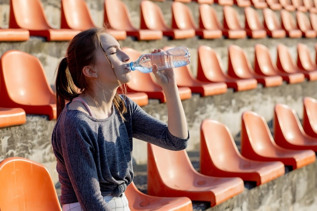 Sportliches, junges, attraktives Mädchen in Sportbekleidung entspannt sich nach hartem Training, sitzt und trinkt Wasser aus einer speziellen Sportflasche, nachdem sie auf einem Stadion gelaufen ist