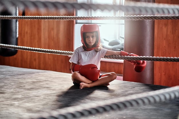 Sportliches junges Mädchen sitzt auf sonnigem Ring und trägt ihre Ausrüstung - Helm und Boxhandschuhe.