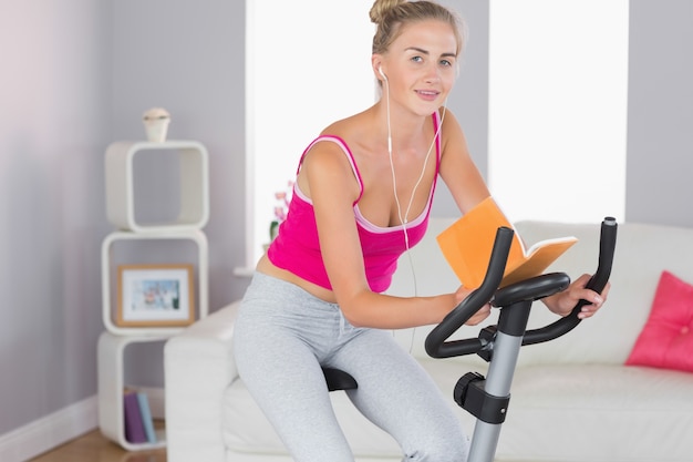 Sportliches lächelndes blondes Training auf dem Hometrainer, der ein Buch liest