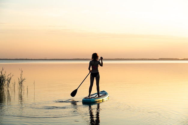 Sportliches Mädchen an Bord für Rotzsurfen. Eine junge schlanke Frau schwimmt auf einem Brett mit einem Paddel. Surfen. Sommerspaß auf dem Wasser. Gesunder Lebensstil. Frau steht beim Stand-Up-Paddle-Boarding (SUP) auf ruhigem Wasser