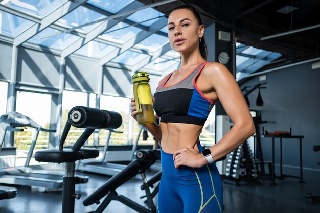 Sportliches Mädchen, das im Fitnessstudio mit Wasserflasche steht