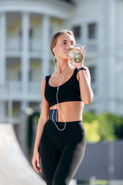 Sportliches Mädchen in der schwarzen Sportkleidung mit Kopfhörern trinkt Wasser.