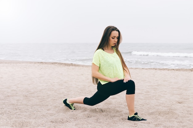 Sportliches schlankes Mädchen, das das Ausdehnen auf den Strand an einem bewölkten Tag tut