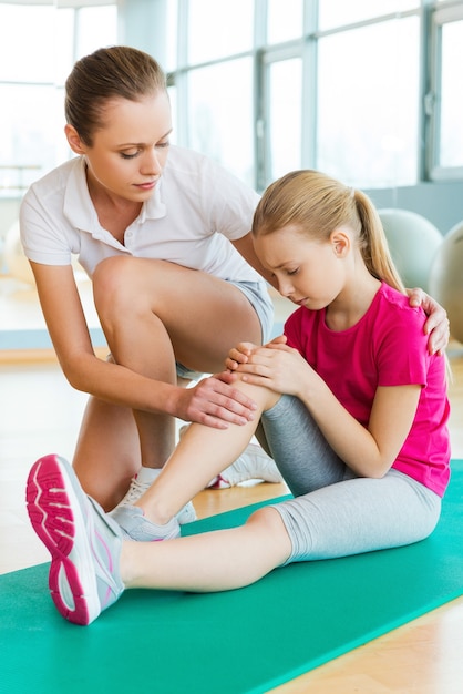 Sportverletzung. Frustriertes kleines Mädchen, das auf Gymnastikmatte sitzt und ihr verletztes Bein berührt, während junge Frau sie tröstet