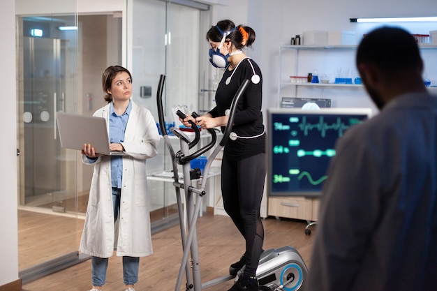 Sportwissenschaftler, der die Herzfrequenz des Athleten überwacht, während eine Frau mit Maske auf einem Crosstrainer läuft und mit einem Arzt spricht. Arzt mit Laptop zur Kontrolle von EKG-Daten im modernen Labor