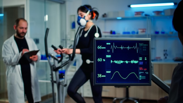 Sportwissenschaftler, der die Herzfrequenz eines Athleten überwacht, während eine Frau mit Maske auf einem Crosstrainer läuft und mit einem Arzt spricht