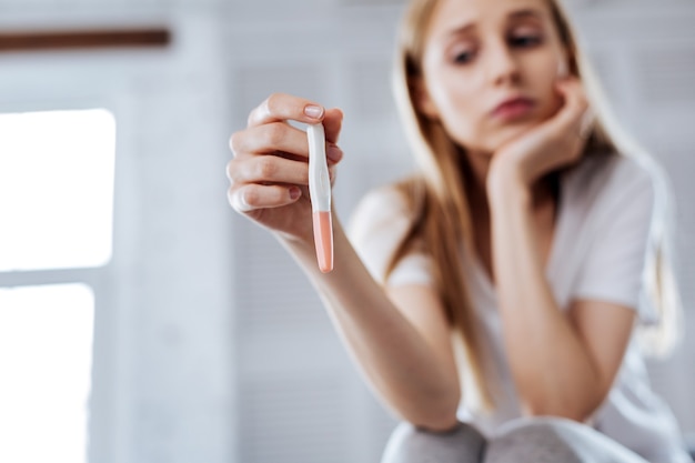 Foto sprachlos. ernsthafte junge frau, die ihren schwangerschaftstest sitzt und hält