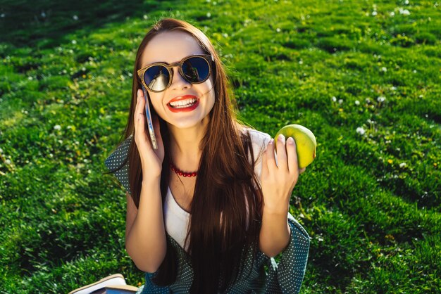 Sprechende Frau hält Handy, sitzt auf Gras im Kleid, Sonnenbrille