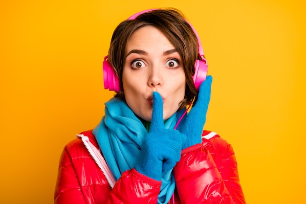 Sprich nicht! Nahaufnahmefoto der schönen Dame hören moderne Kopfhörer halten Finger auf den Lippen tragen stilvollen lässigen roten Mantel blauen Schal Handschuhe Rollkragenpullover