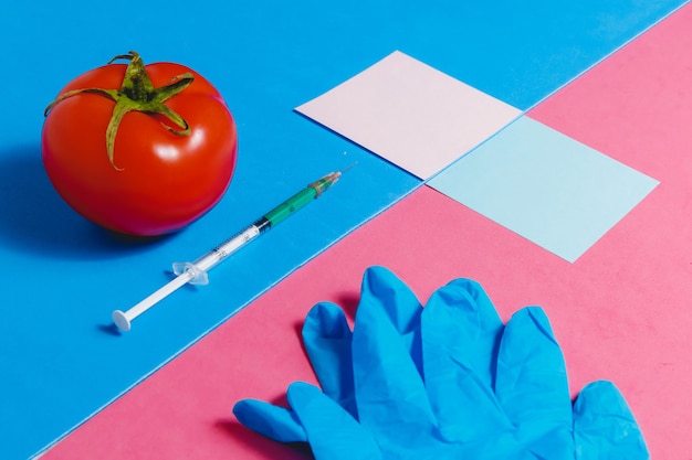 Spritze, Aufkleber, blaue Handschuhe und rote Tomate auf rosa und blauem Hintergrund,