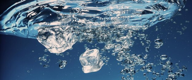 Spritzendes Wasser und Eiswürfel. Eis spritzt in ein Glas Wasser. Unter Wasser fallen strömende Eiswürfel in einen klaren Bewässerungshintergrund. Erfrischendes, kühles Trinken. Eis in einem Glas mit blauem Hintergrund