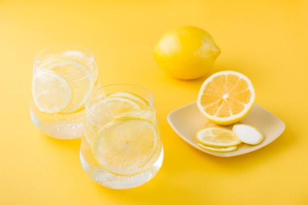 Sprudelwasser mit Zitrone und Eis in Gläsern und Zitronenscheiben auf einer Untertasse auf einem gelben Tisch.