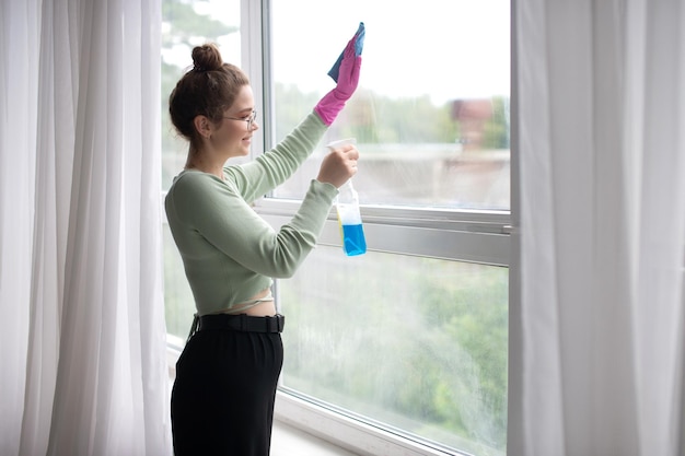 Sprühen Sie das Spray, um das Fenster zu reinigen Glückliche junge Frau, die die Reinigung durchführt