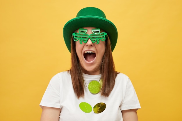 St. Patrick's Day Party Extrem glücklich überglücklich Frau mit grünem Hut und Kleebläsern steht isoliert über gelbem Hintergrund schreit von Aufregung freut sich