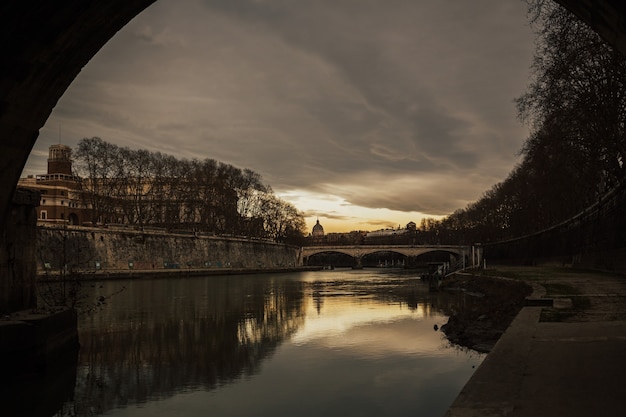 Stadtbild und Panoramablick der alten Brücke mit warmen Sonnenuntergangshimmelreflexionen im Wasser des Tiberflusses und Kuppel der St. Peters Kathedrale