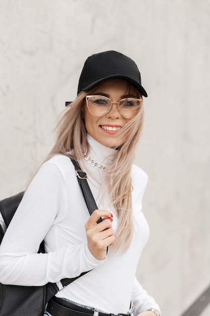 Stadtporträt eines schönen, modischen, glücklichen Mädchens mit einem Lächeln und einer Brille in einem modischen weißen Pullover und einer Mütze mit einem Rucksack in der Stadt in der Nähe einer grauen Betonwand