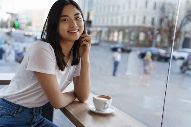 Städtische junge Frau, die auf Handy spricht, im Café nahe Fenster sitzt und lächelt, Tasse Kaffee an warmem Sommertag trinkend