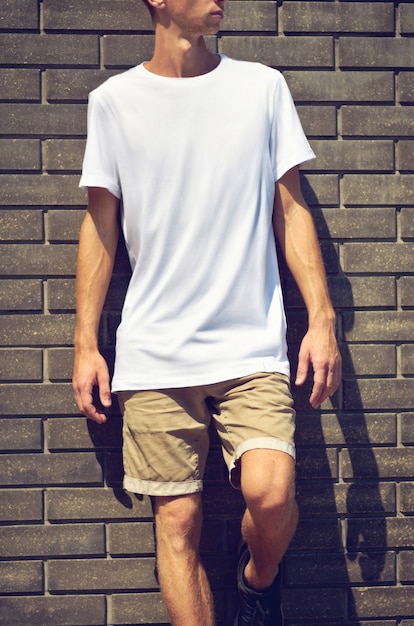 Städtisches Modell von Kleidung. Junger Mann steht auf einem Backsteinmauerhintergrund in einem weißen T-Shirt und braunen Shorts. Vorlage bereit für Ihr Design