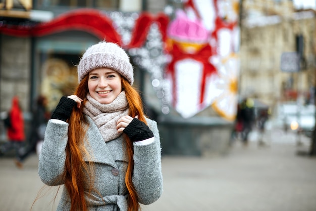 Städtisches Porträt einer entzückenden Ingwerfrau mit langen Haaren, die warme Kleidung trägt, die in der Stadt spazieren geht. Platz für Text