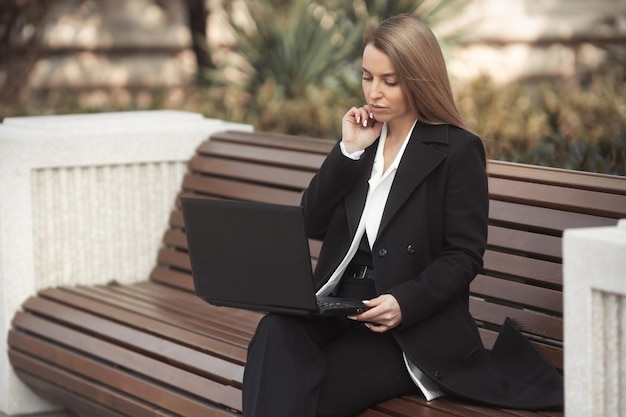 Städtisches Porträt einer schönen jungen Geschäftsfrau, die aus der Ferne mit Laptop arbeitet und telefoniert