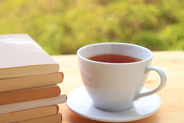 Stapel Bücher und eine Tasse heißen Tee auf einem Tisch am Fenster mit unscharfem Hintergrund des Herbstlaubs.