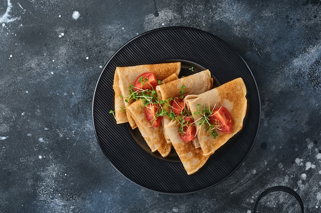 Stapel glutenfreier Buchweizenmehl-Crepes-Pfannkuchen mit Kirschtomaten und Rucola-Mikrogrün auf schwarzem Teller auf dunklem Hintergrund, hausgemachtes gesundes Backen zum Frühstück. Attrappe, Lehrmodell, Simulation. Platz kopieren.