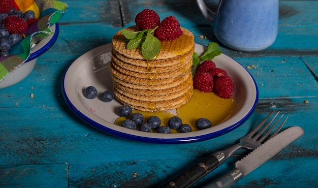 Stapel holländischer Karamellwaffeln mit Himbeeren, Blaubeeren und Honig auf blauem hölzernem Hintergrund.