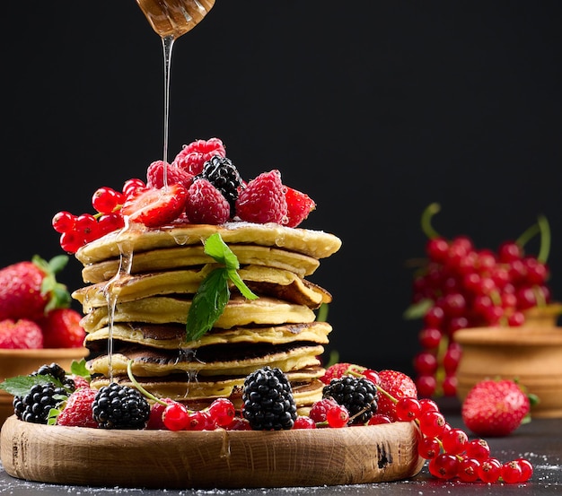 Stapel Pfannkuchen mit frischem Obst, bestreut mit Honig auf schwarzem Hintergrund