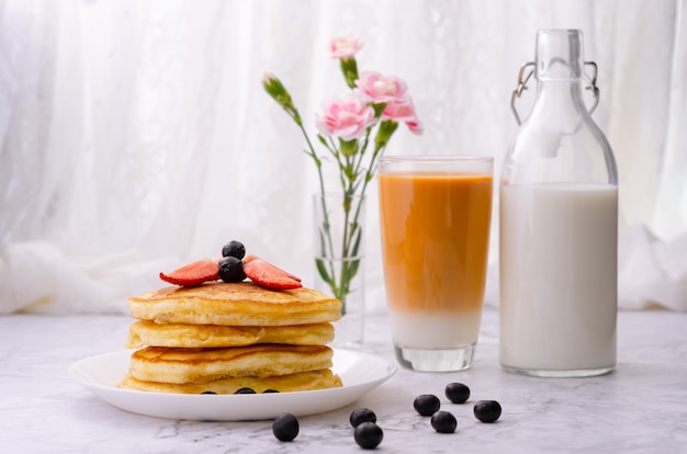 Stapel Pfannkuchen mit frischen Erdbeeren und Blaubeeren auf einem weißen Teller, eine Flasche Milch, ein Glas Milchtee und eine Vase mit rosa Blumen auf einem Marmortisch