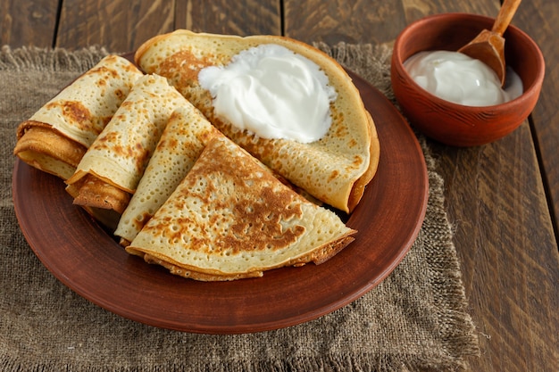 Stapel Pfannkuchen oder Crpes auf einem Teller mit Sauerrahm serviert. Traditionelles Essen für die russische Pfannkuchenwoche. Hölzerner Hintergrund.