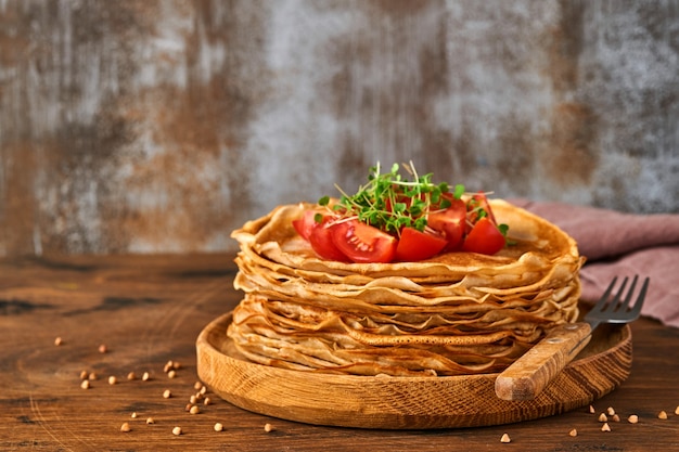 Stapel von glutenfreien Buchweizenmehl-Crepes-Pfannkuchen mit Kirschtomaten und Rucola-Mikrogrün auf Holzplatte, hausgemachtes gesundes Backen zum Frühstück.