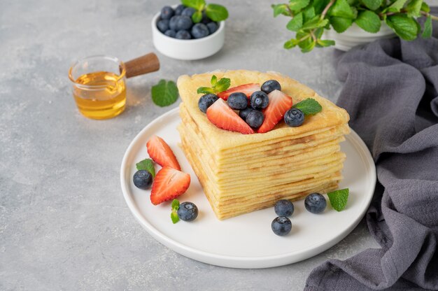 Stapel von hausgemachten Crepes serviert mit frischen Blaubeeren und Erdbeeren auf einem weißen Teller auf einem grauen Betonhintergrund. Essen für Maslenitsa. Speicherplatz kopieren.