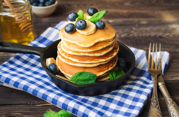 Stapel von hausgemachten Pfannkuchen in Eisenpfanne mit Blaubeeren, Bananen- und Minzblättern auf rustikalem hölzernem Hintergrund.