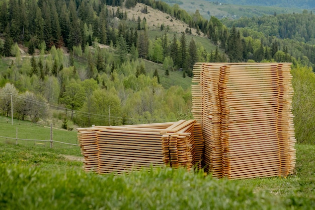 Stapel von Holzbrettern auf grünem Gras mit Bergen im Hintergrund, Beplankung für den Bau. Holzstapel aus Holzrohlingen Baumaterial.