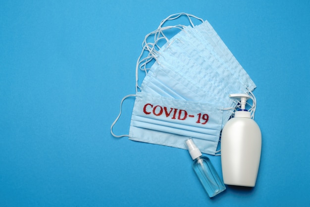 Foto stapel von medizinischen medizinischen einwegmasken mit covid-19-zeichen und alkohol-händedesinfektionsmittel-antiseptikum auf blauem hintergrund