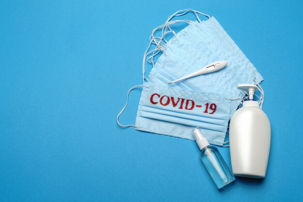 Foto stapel von medizinischen medizinischen einwegmasken mit covid-19-zeichen und alkohol-händedesinfektionsmittel-antiseptikum auf blauem hintergrund