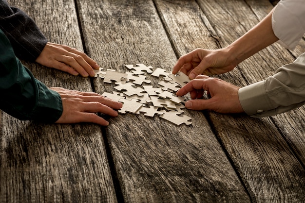 Stapel von Puzzleteilen, die mit vier Händen (männlich und weiblich) auf einem strukturierten Holzschreibtisch liegen und jeweils eine nehmen. Konzeption der Teamarbeit und Strategieplanung.